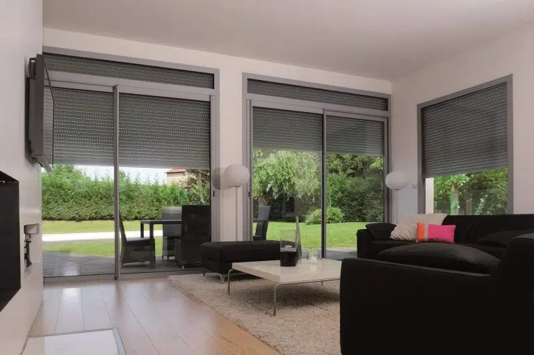 Salon moderne avec larges baies vitrées équipées de volets roulants gris.