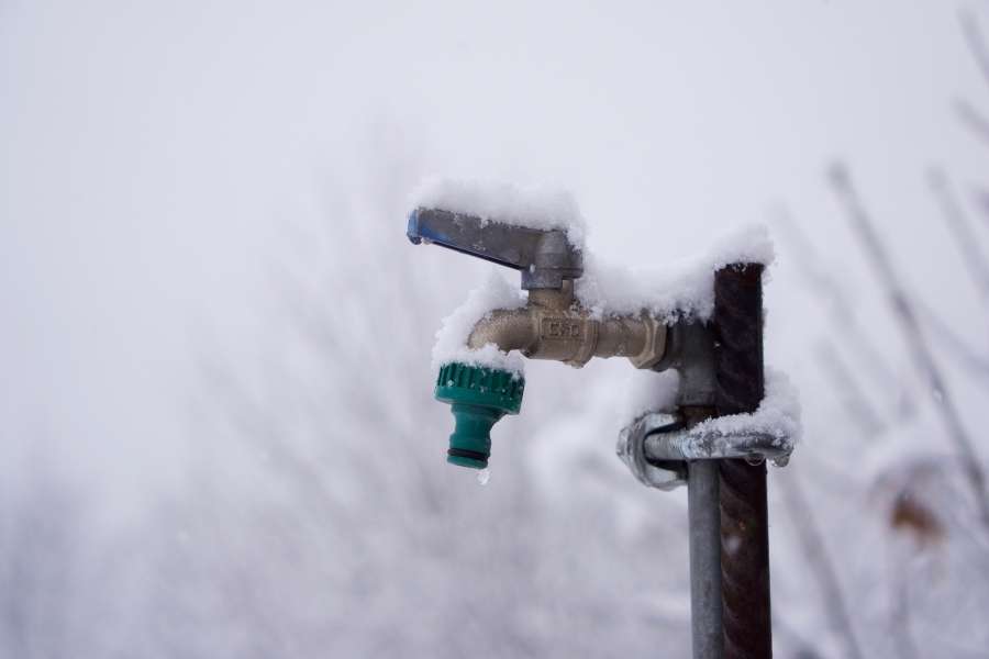 Un robinet de jardin en laiton et son raccord rapide vert sont recouverts d'une épaisse couche de neige fraîche, illustrant le froid intense de l'hiver.