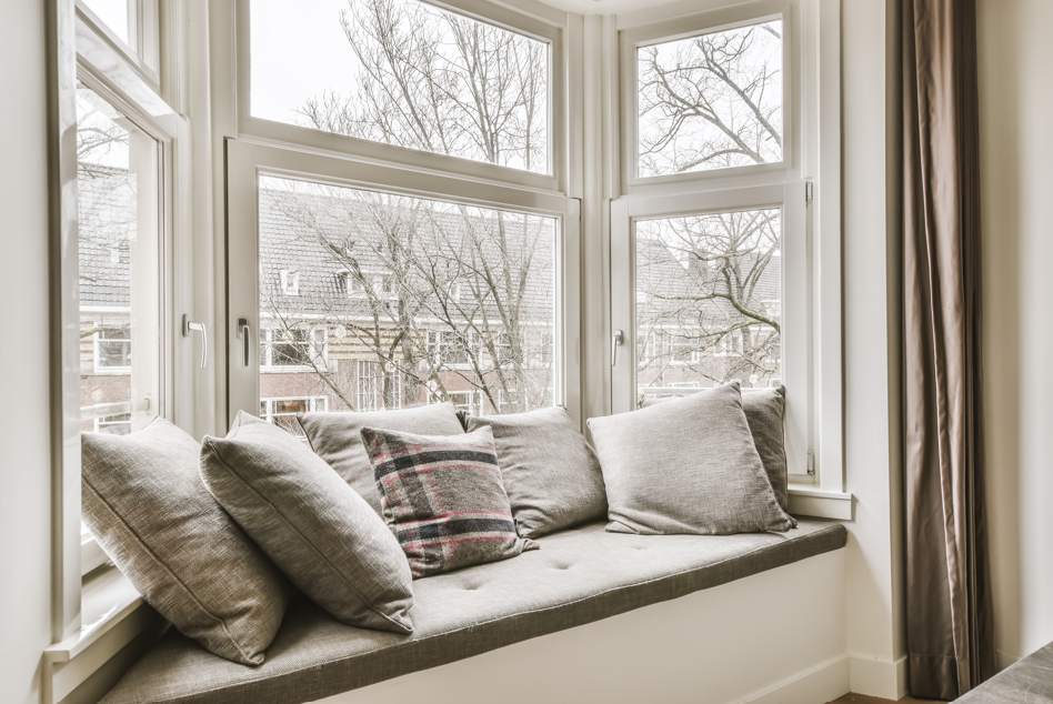Un confortable coin lecture aménagé sur le rebord d'une grande fenêtre de baie en PVC blanc, offrant une vue sur des bâtiments en briques et des arbres nus, parfait après une renovation fenetre menuiserie pvc prime energie.
